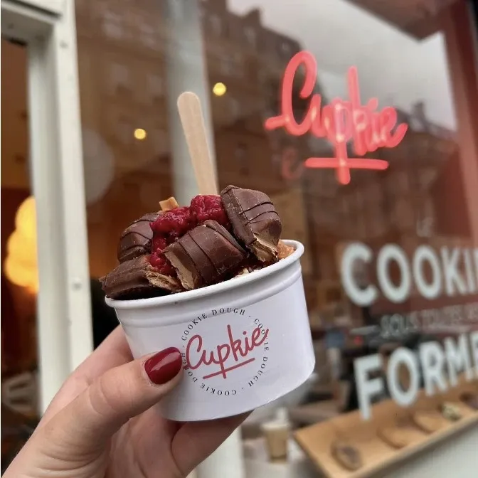 Cup de cookie dough garnie de framboises et de chocolat, tenue devant la vitrine Cupkie